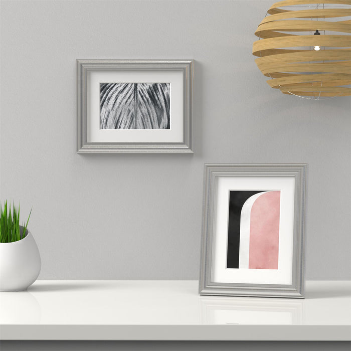Silver picture frame with white mount displaying art on a white shelf against a grey wall.