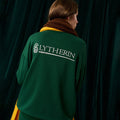 Person wearing a green sweater with 'SLYTHERIN' text and logo on the back against a dark curtain.