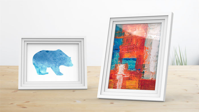 White picture frame displaying a blue bear silhouette and a colorful abstract painting on a wooden table.